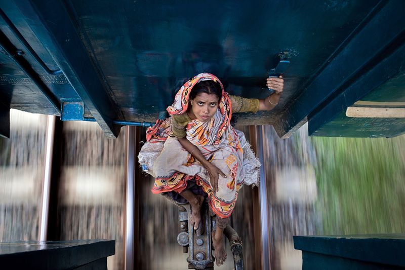 Опасное путешествие. Женщина едет между вагонами поезда, направляющегося к северу от Дакки – столицы Бангладеш. Ее багаж лежит под вагоном перед ней. Фото сделано в месяц Рамадан, кульминацией которого является Ураза-байрам. Десятки тысяч людей покидают город и отправляются в родные деревни, чтобы отметить праздник с семьями. Поезда переполнены, и многие из тех, кто не успел купить билеты или не смогли позволить себе купить их, едут на крышах или между вагонами, как эта женщина. (Photo and caption by Amy Helene Johansson)